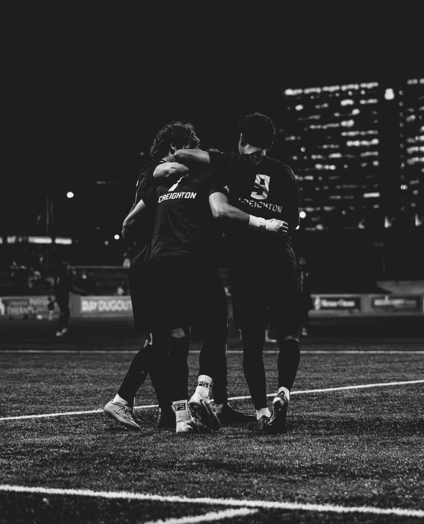 Two Creighton players in dark kits embrace on the field at night.