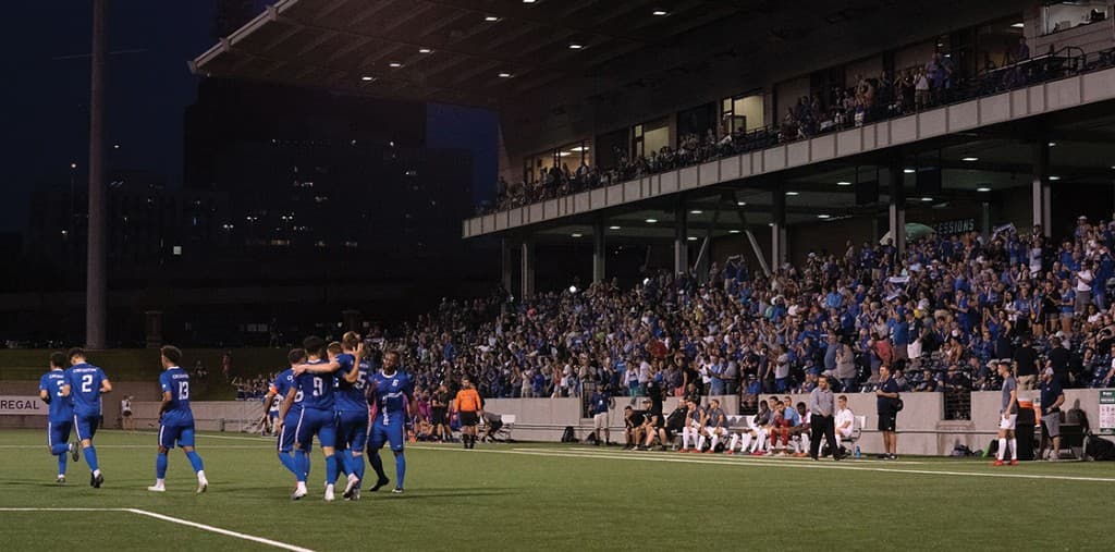 Creighton players in blue celebrate on the field at night while the stands stay packed.